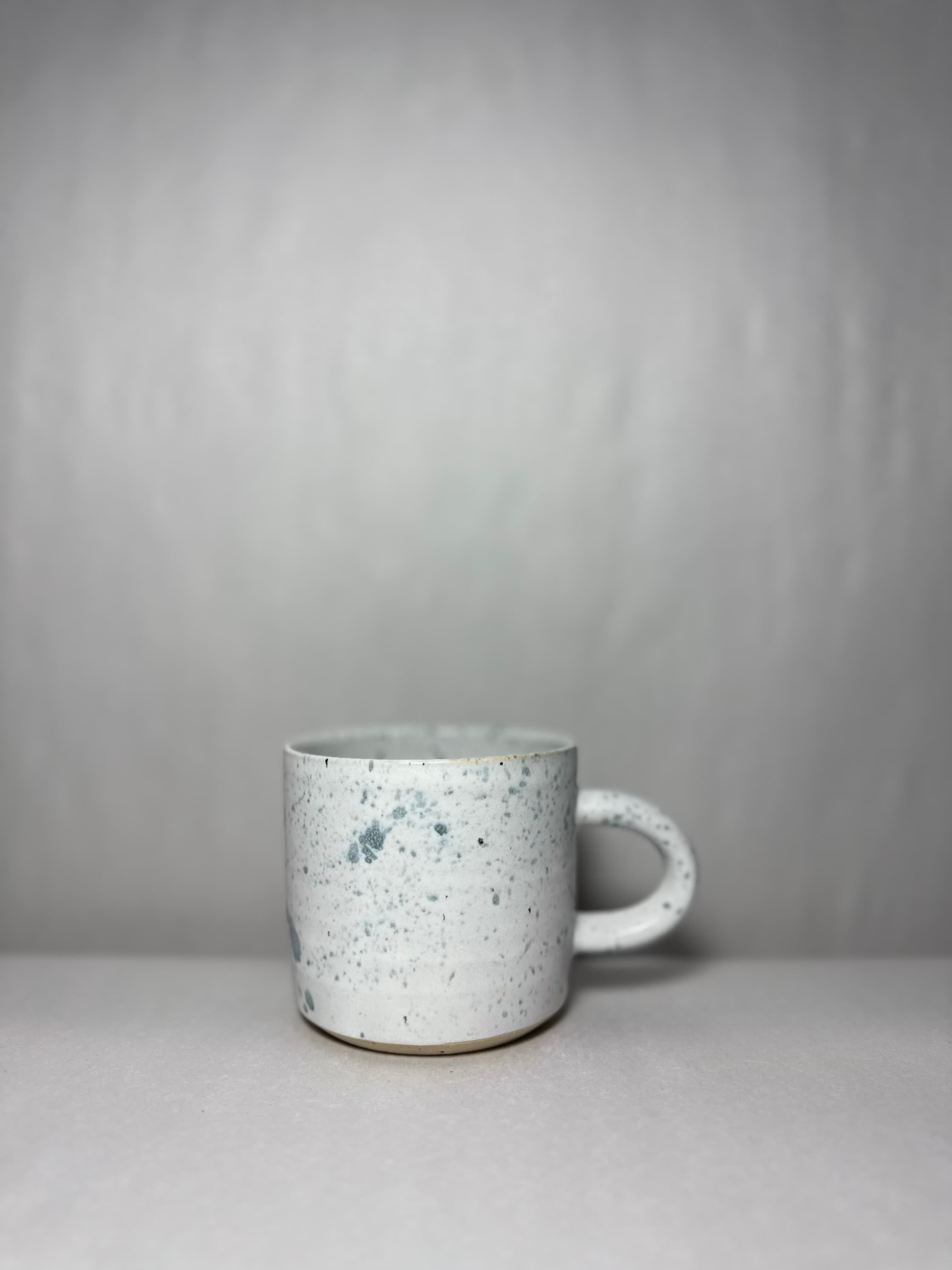 A single speckled white mug photographed against a soft gray ground